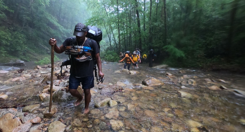 a person carrying a backpacking uses a walking stick to cross a creek. there are green trees in the background.